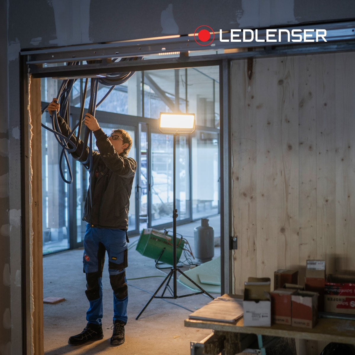 Electrician pulling cable through a ceiling on a fit-out site, lit by a Ledlenser floodlight on a tripod