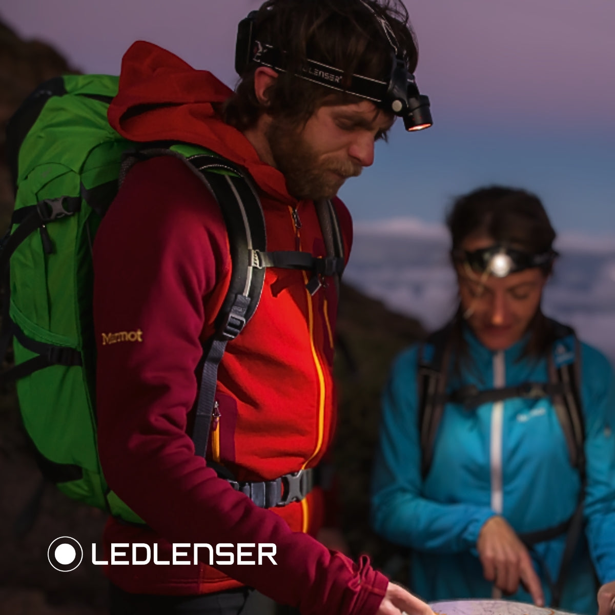 Two hikers with rucksacks reading a map at dusk under Ledlenser head torches