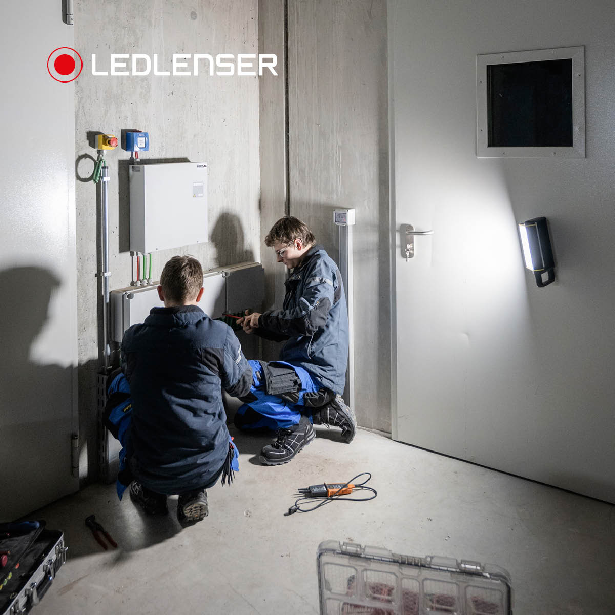 Two electricians kneeling at a wall-mounted distribution box, lit by a Ledlenser area light fixed to a steel door