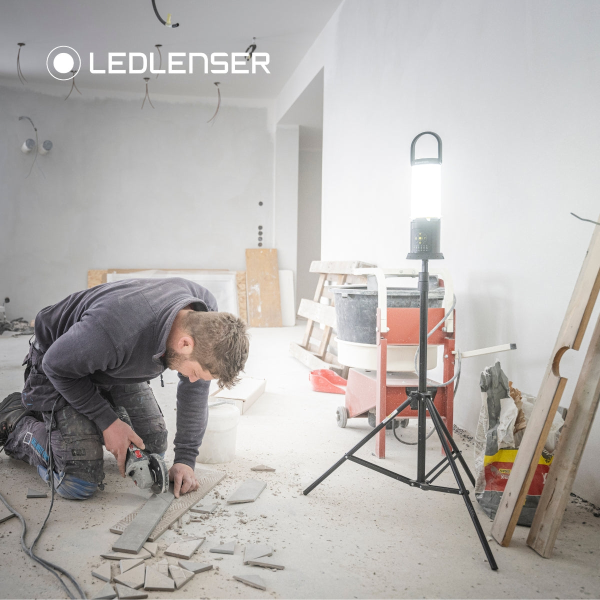 Tradesman cutting tile with an angle grinder in a stripped-out room, lit by a Ledlenser AL10R Work on a tripod