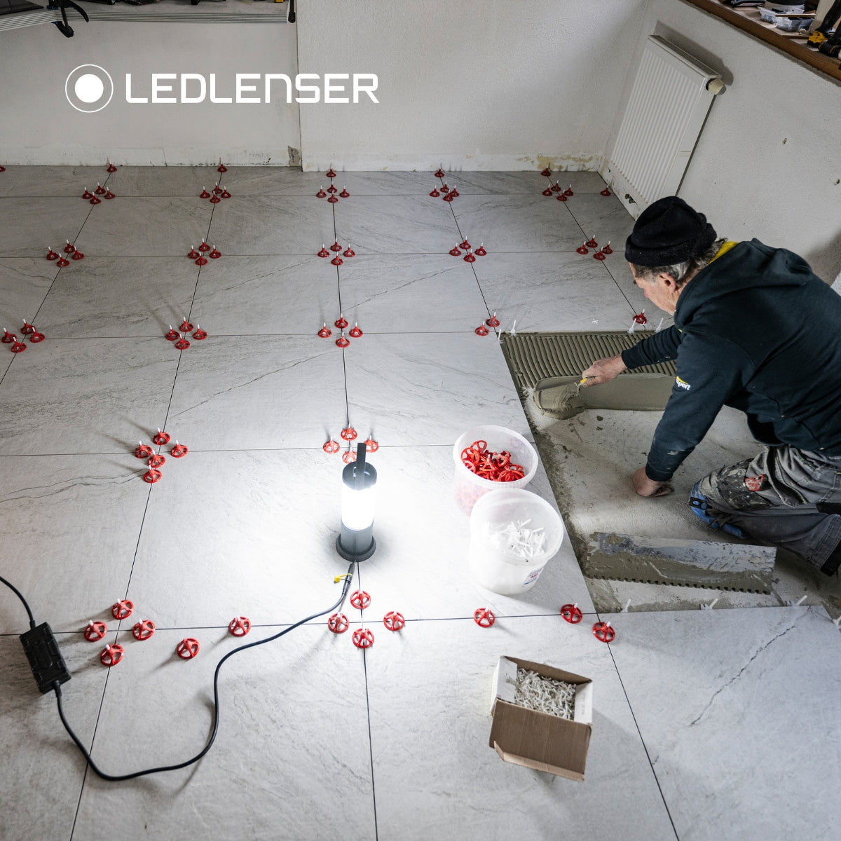 Overhead view of a tiler spreading adhesive across a large floor lit all round by a mains-connected Ledlenser AL10R Work