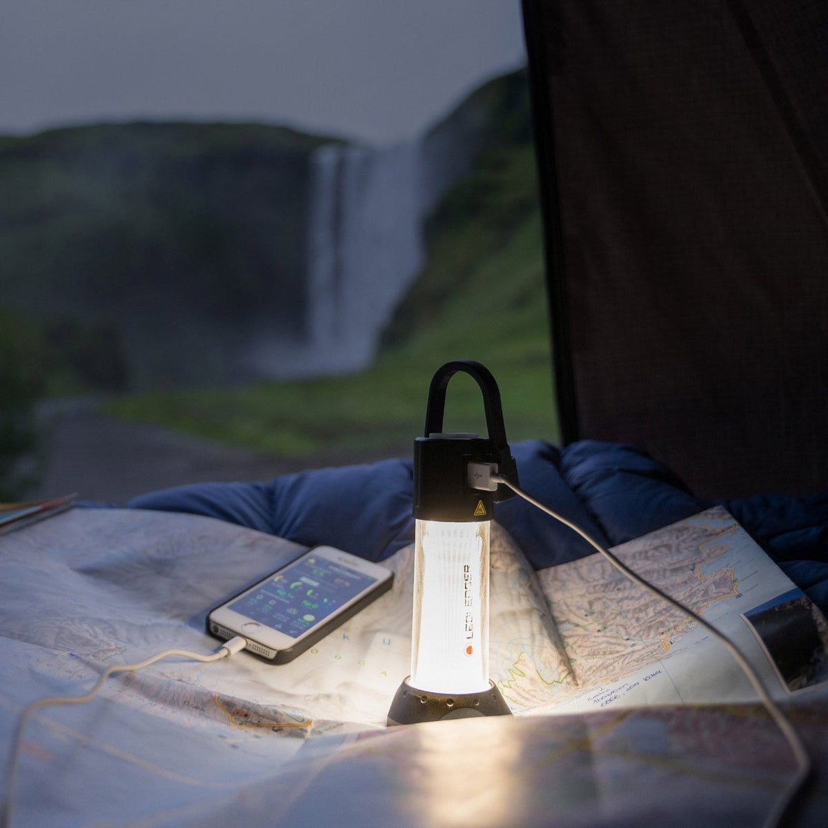 Ledlenser ML6 charging a phone in a tent beside a map, with a waterfall outside