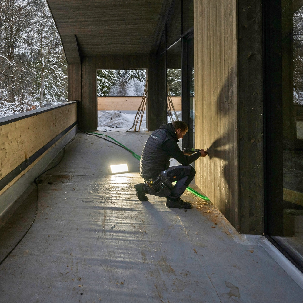 Tradesman sealing a timber wall on a covered balcony, lit by a Ledlenser floodlight standing on the floor