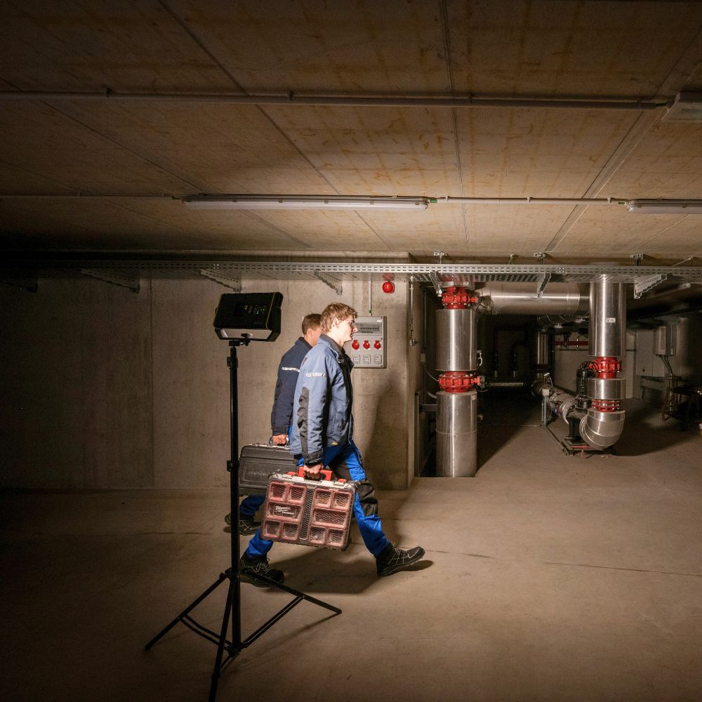 Two engineers carrying toolboxes through a basement plant room lit by a Ledlenser area light on a tripod