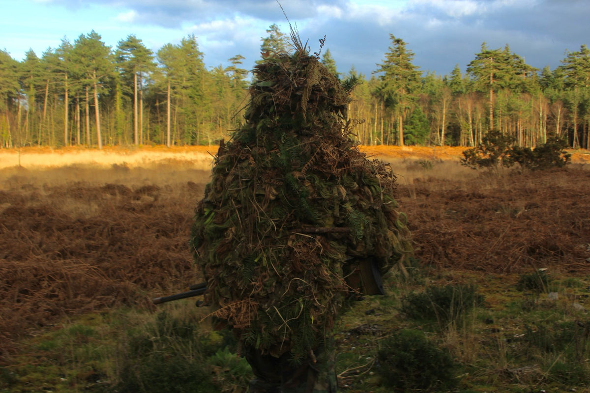 Rear view of the full cape ghillie suit dressed with bracken and conifer, standing in open heathland below a treeline