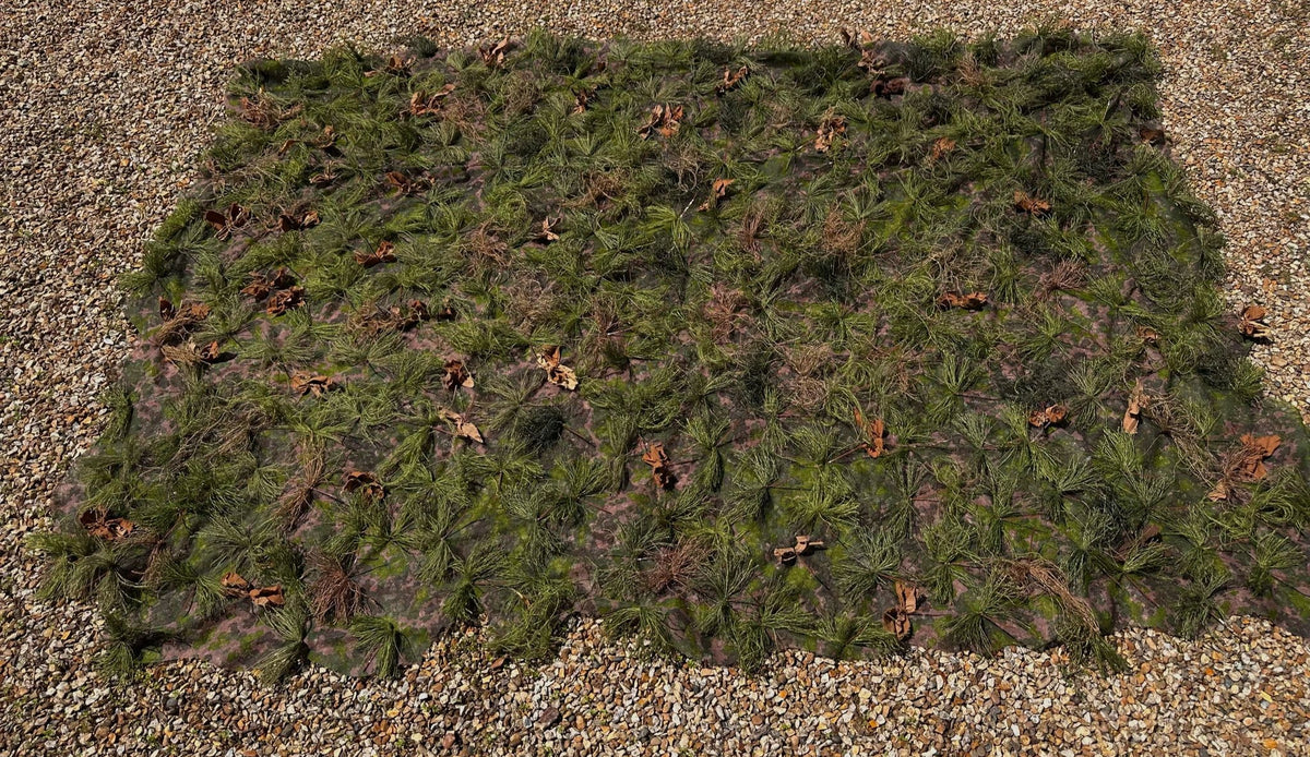 Overhead view of the Zero Alpha Designs hide sheet spread flat on gravel, garnish carried right out to the edges of the camouflage base