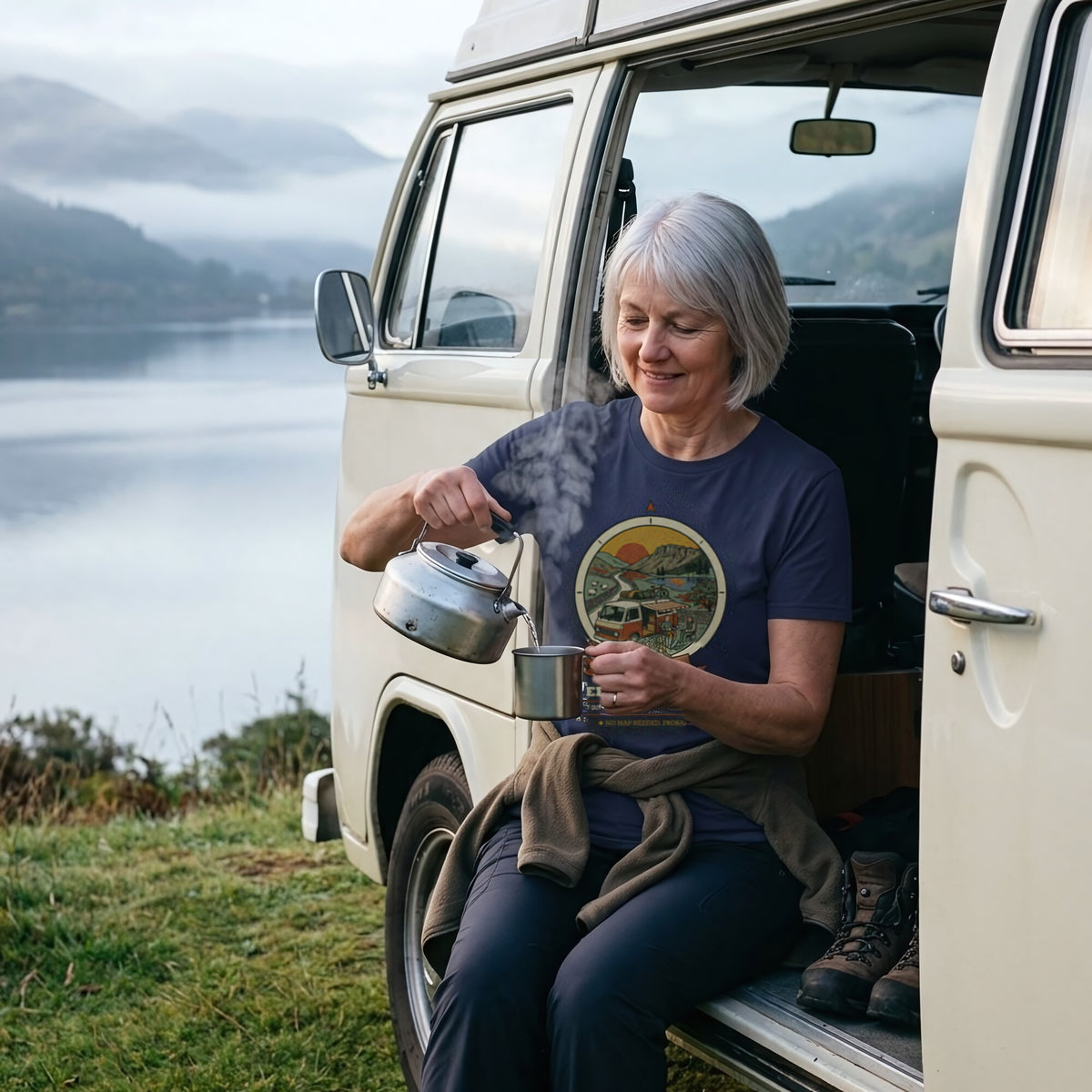 Woman in the Adventure Before Dementia Campervan T-Shirt in Navy pouring tea from a kettle in the campervan doorway above a misty loch