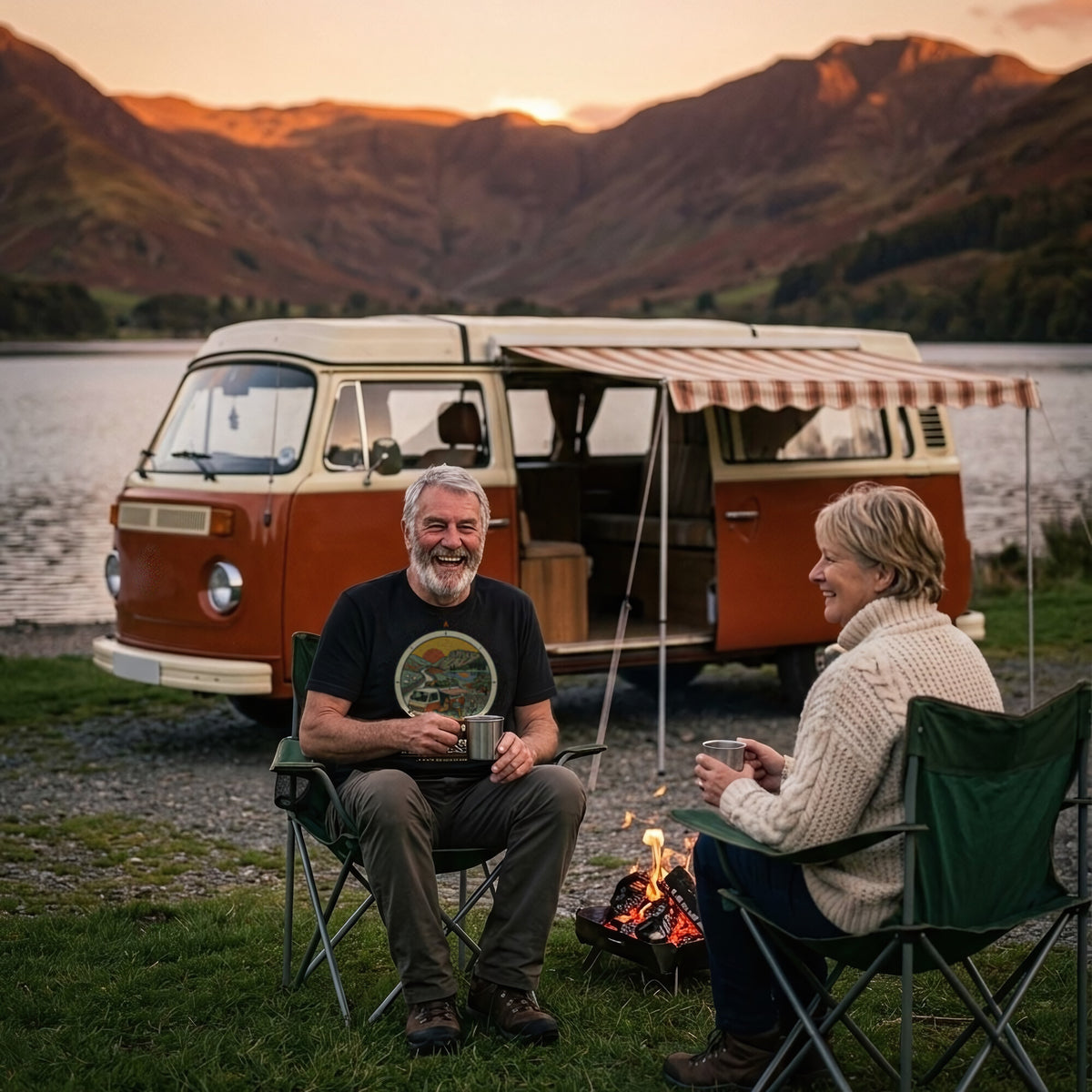 Man in the Adventure Before Dementia Campervan T-Shirt in Vintage Black laughing with his wife in camping chairs by a campfire, their campervan parked by a lake at dusk
