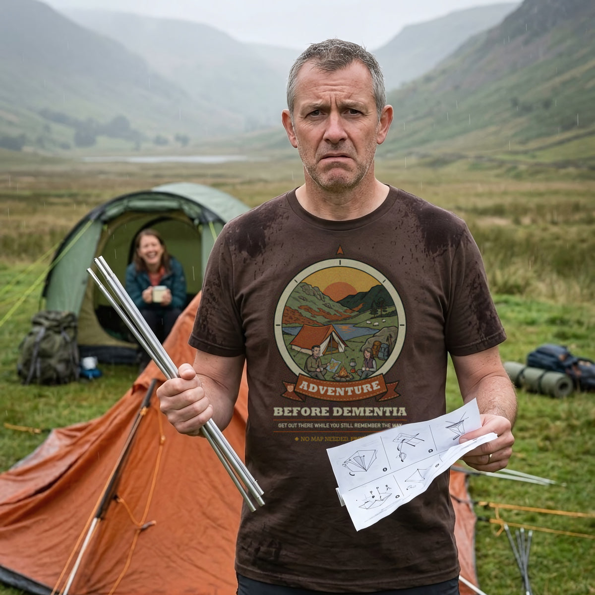 Man in the Adventure Before Dementia Camping T-Shirt in Brown puzzling over tent instructions in the rain