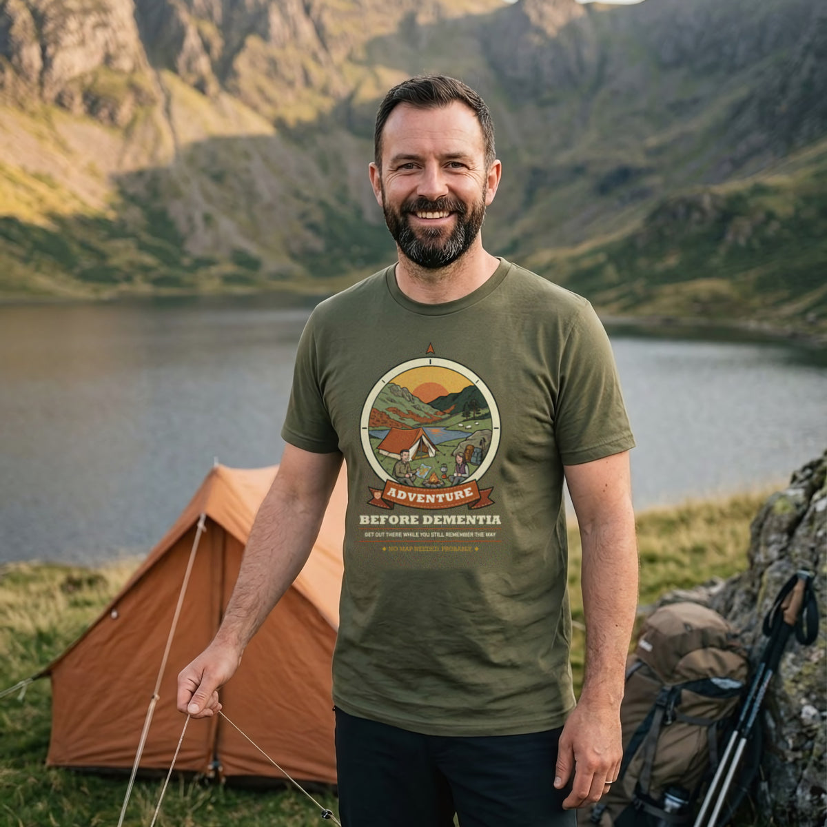 Man wearing the Adventure Before Dementia Camping T-Shirt in Military Green beside an orange ridge tent above a mountain tarn