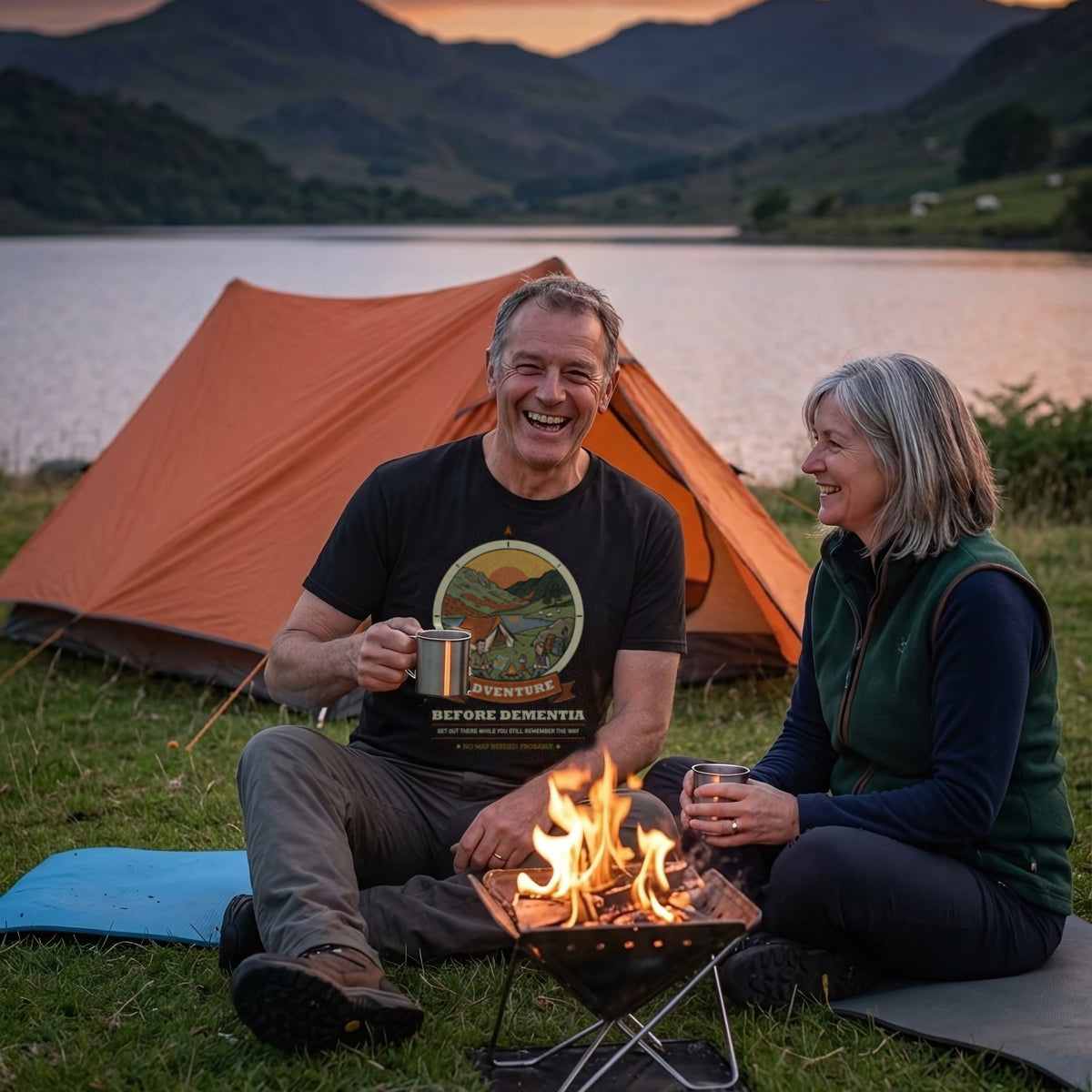 Couple by a campfire at a lakeside pitch at dusk, the man in the Adventure Before Dementia Camping T-Shirt in Vintage Black