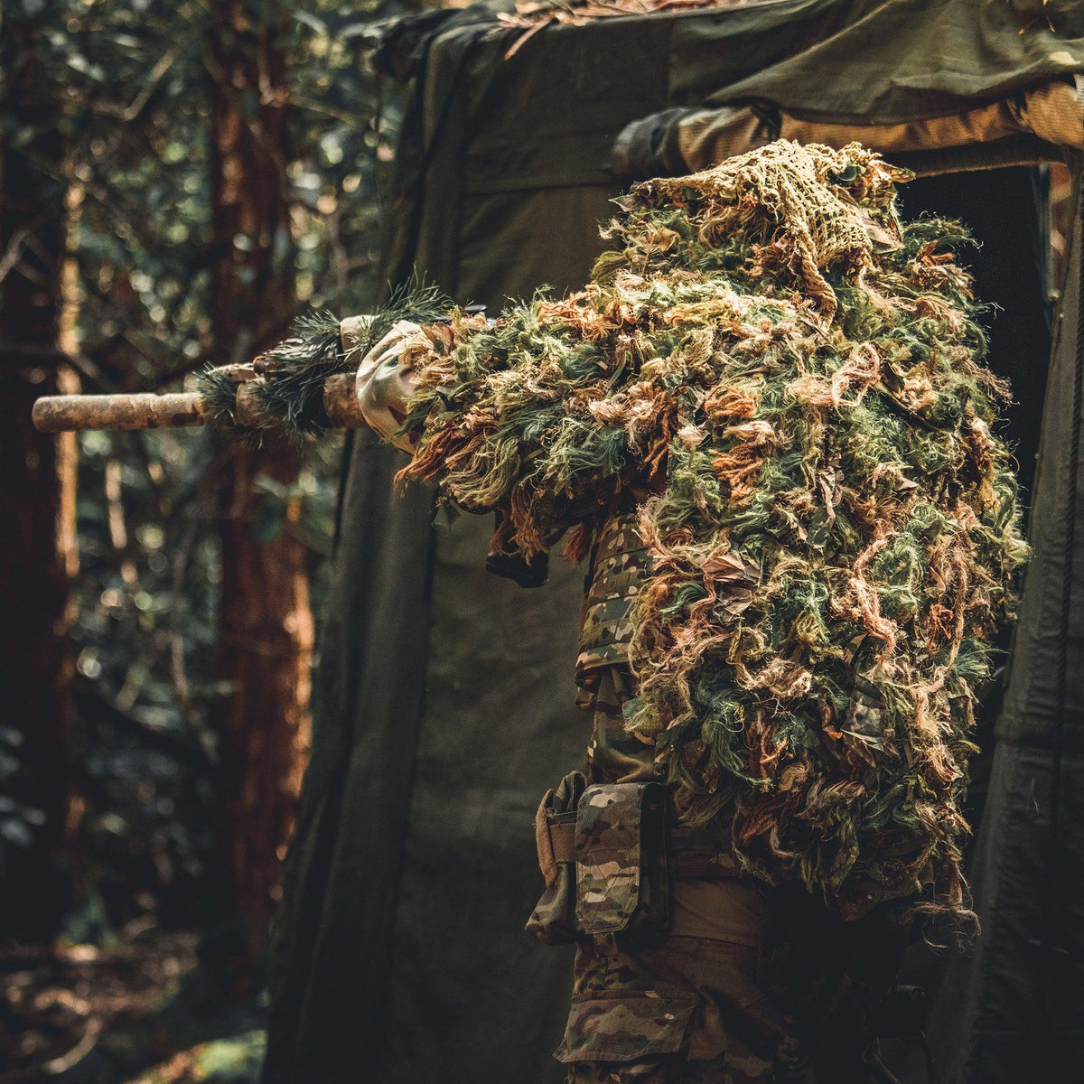 Shooter in a Zero Alpha Designs full cape ghillie suit shouldering a wrapped rifle from the mouth of a woodland hide