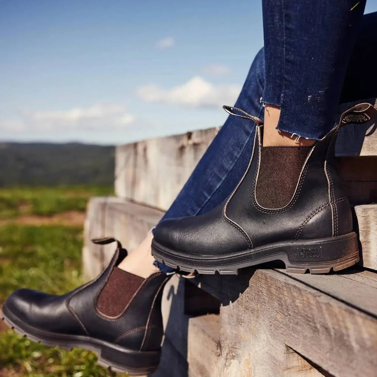 Person seated on timber sleepers wearing brown Mongrel K9 dealer boots, green countryside behind