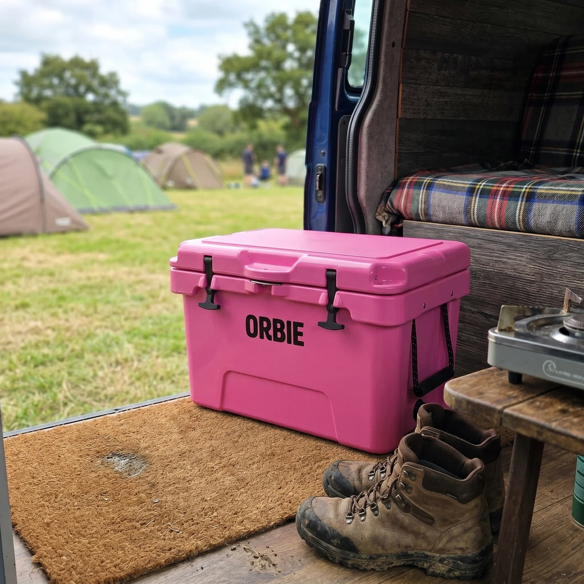 Pink Orbie 35 cool box on the back of a camper van at a campsite, beside walking boots and a camping stove