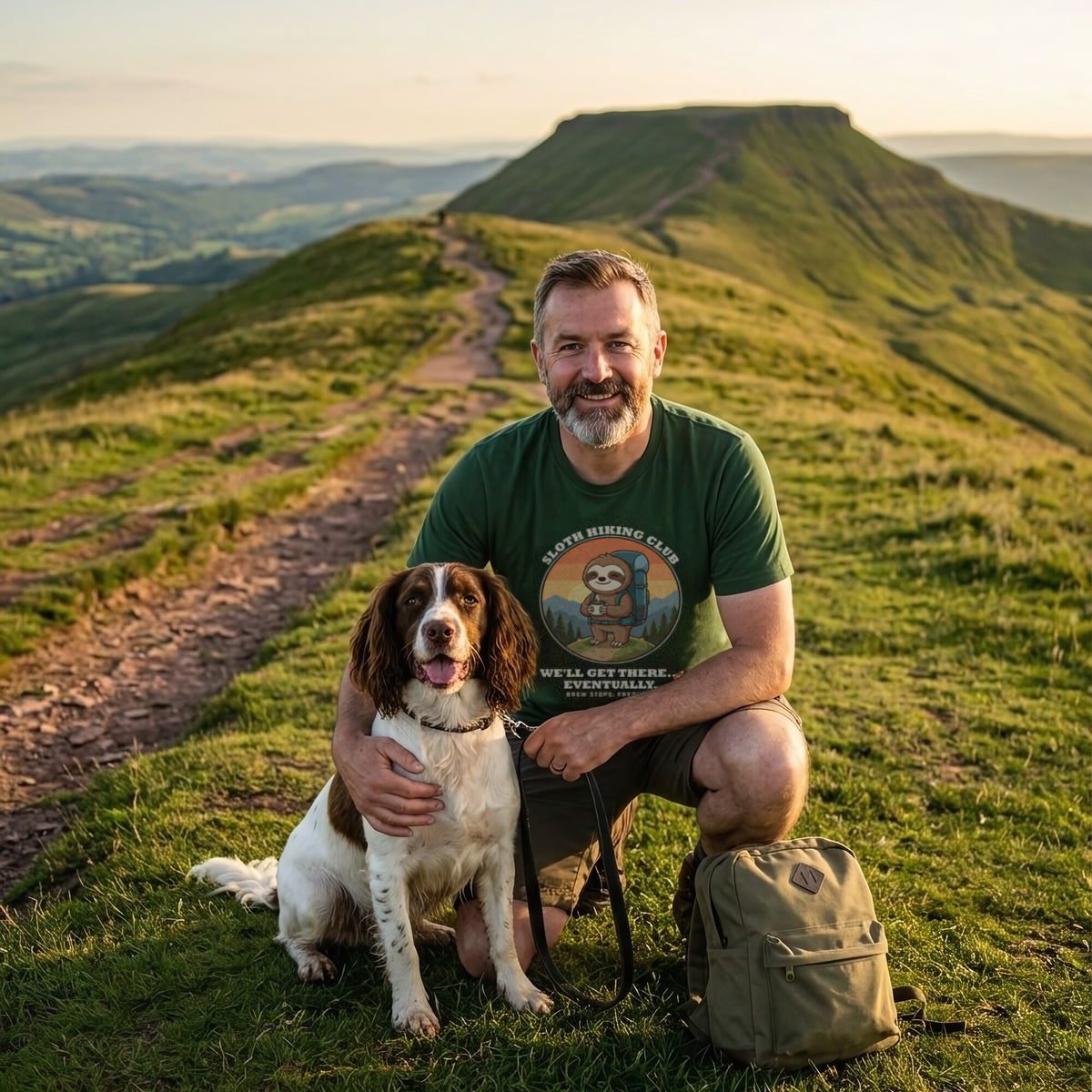 Man crouching with a springer spaniel on a grassy ridge path, wearing the Sloth Hiking Club: Hilltop T-Shirt in Forest