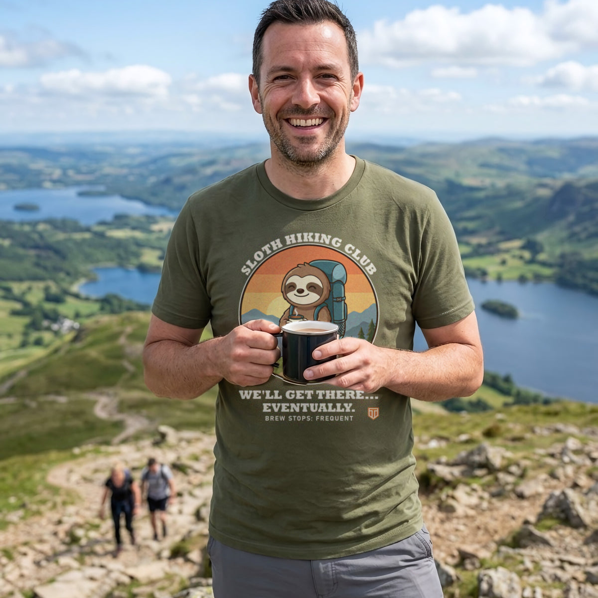 Man with a mug of tea on a Lake District fell top while his friends climb behind, wearing the Sloth Hiking Club: Hilltop T-Shirt in Military Green