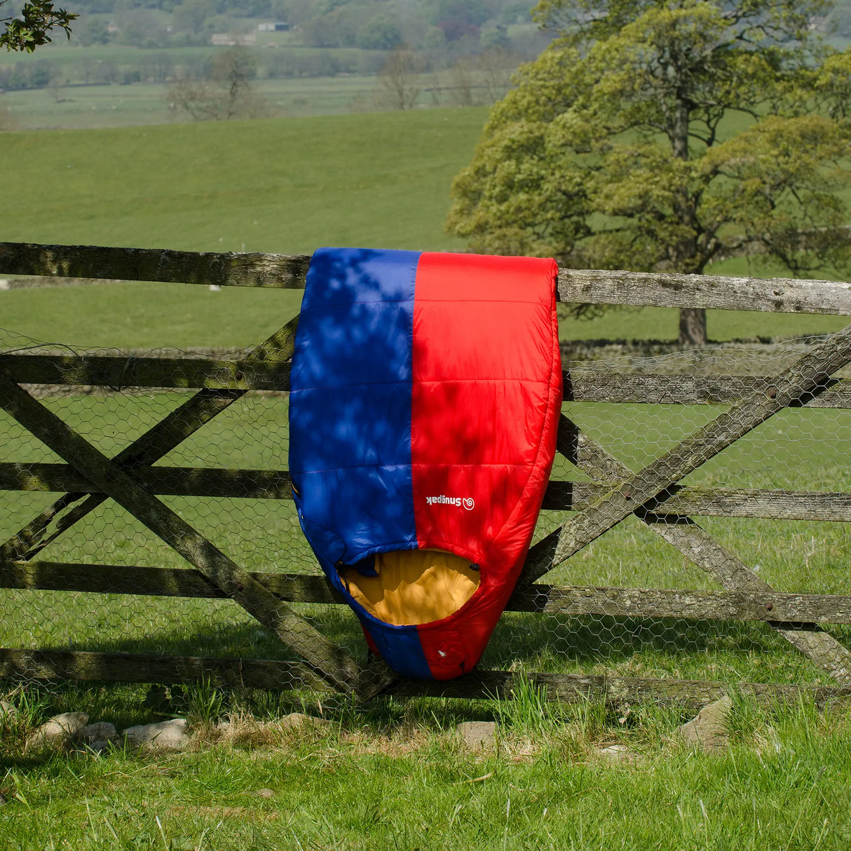 Snugpak Softie Expansion 2 WGTE Sleeping Bag in red and blue draped over a wooden gate in a countryside field
