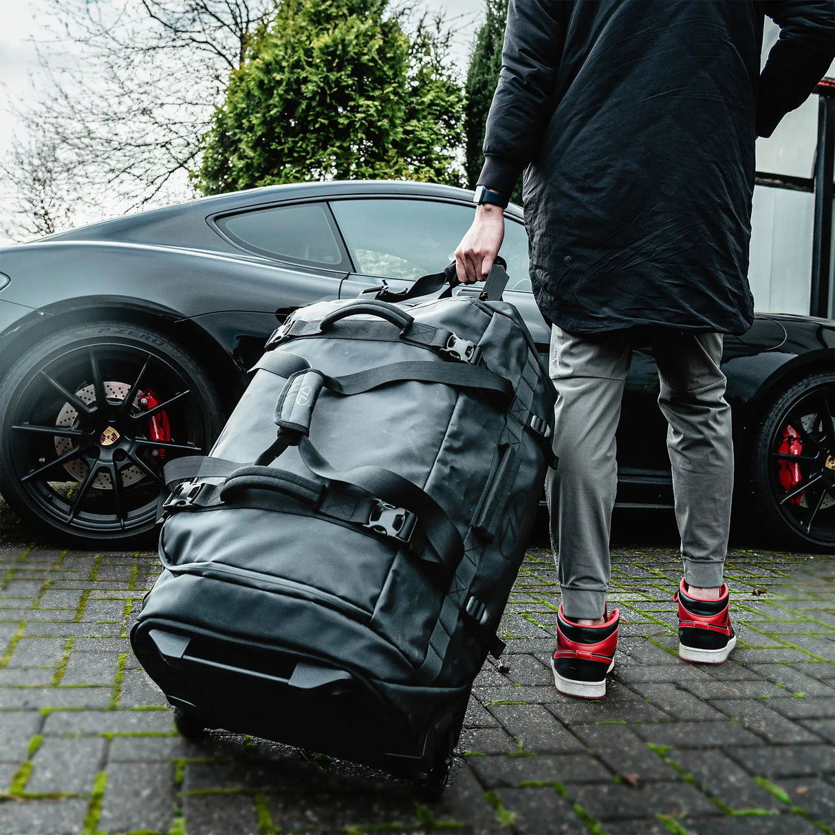 Highlander The Trailer Duffle with Wheels 100L - Black - being used outdoors by a gentleman walking towards a car