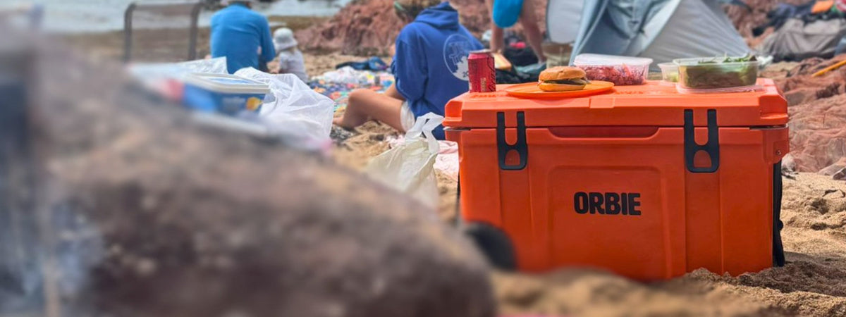 Orange Orbie 35 cool box on a sandy beach with a burger and snacks on the lid, a family picnic behind