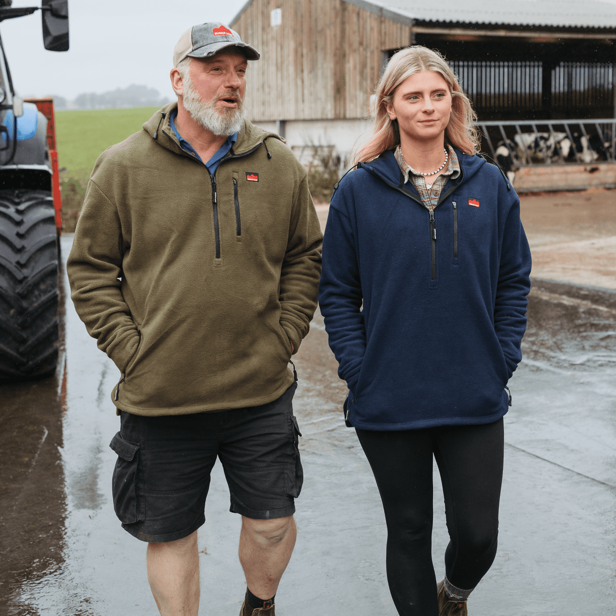 Two people wearing Sugar Loaf Shepherd Polar Fleece Hoodie in Khaki standing on a farm with a tractor and barn in the background.