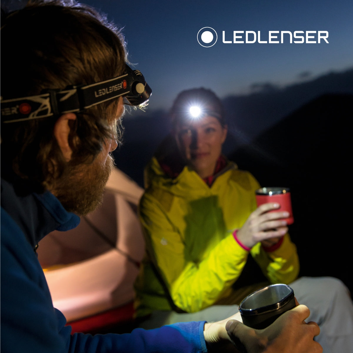 Two campers sharing hot drinks at night, lit by Ledlenser head torches