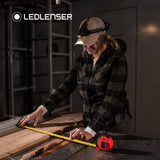 Woman in a cap measuring timber on a saw bench, wearing a Ledlenser head torch