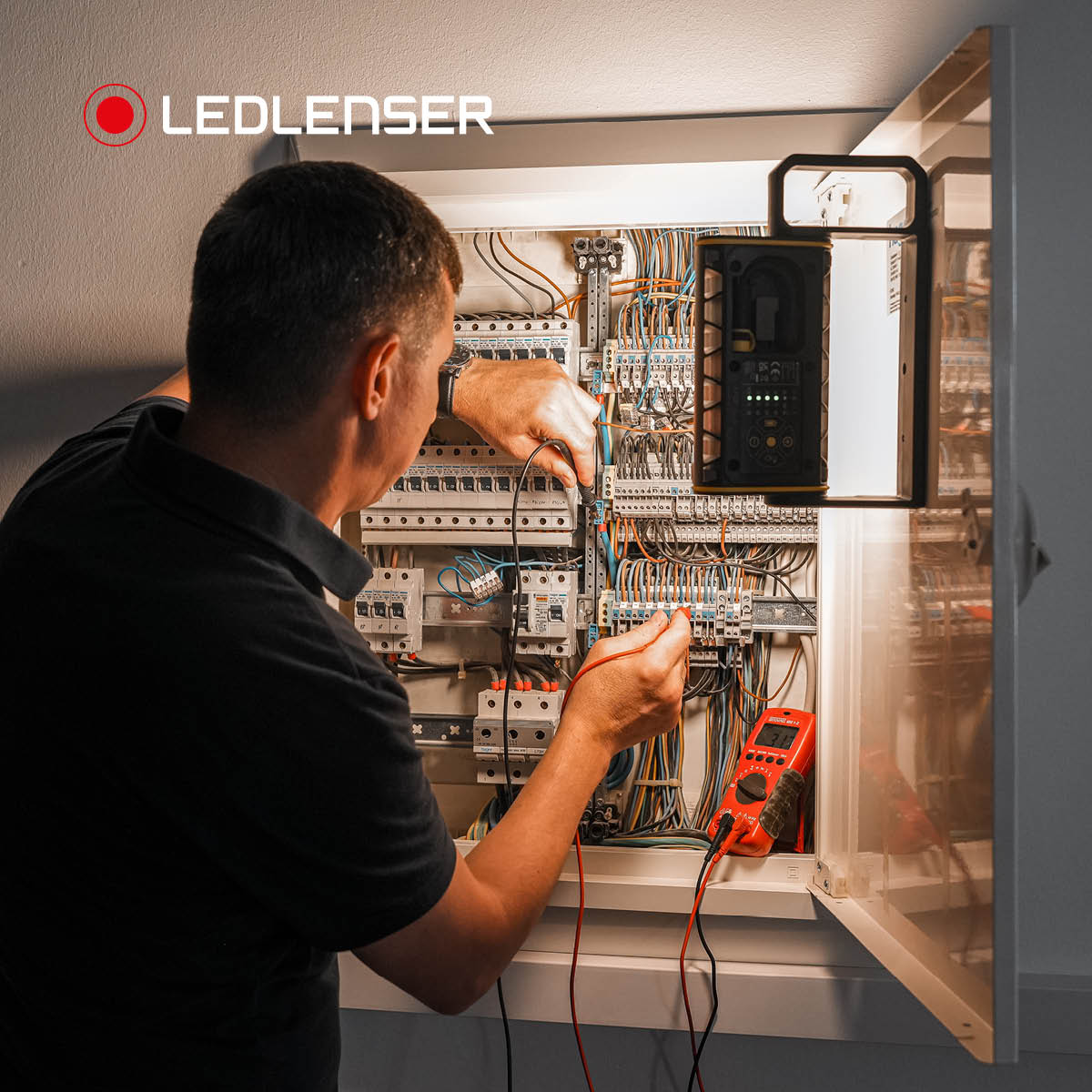 Electrician testing a fuse board with a multimeter, lit by a Ledlenser AF4R Work hung on the cabinet door
