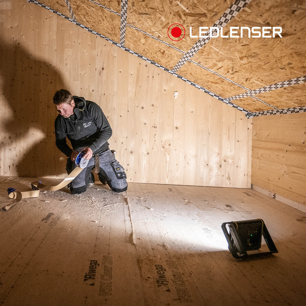 Roofer taping a membrane in a timber loft space, lit by a Ledlenser AF8R Work standing on the floor