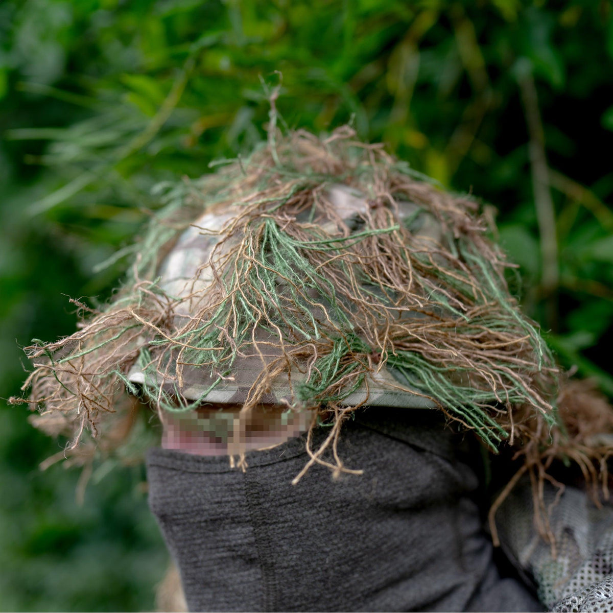 Zero Alpha Designs ghillie boonie hat worn against green foliage, tan and green hessian tied over a camouflage boonie and hanging past the brim over the eyes