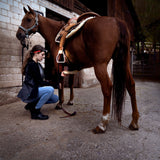 Rider crouching beside a saddled horse in a stable yard, wearing a Ledlenser head torch