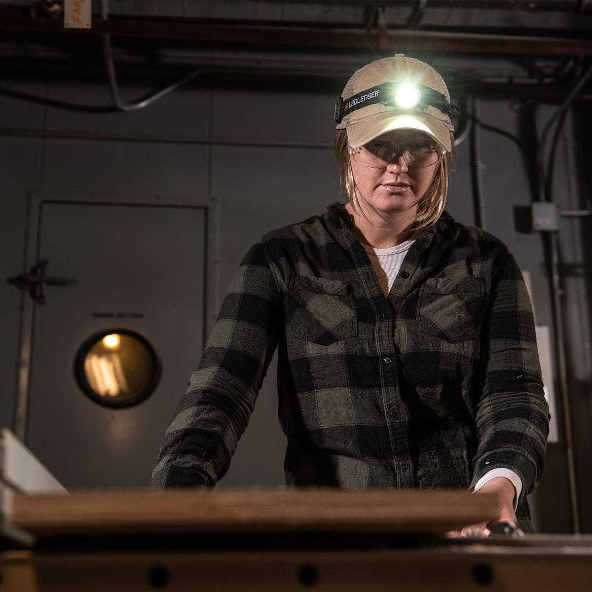 Woman in a cap and safety glasses at a workbench, lit by a Ledlenser head torch