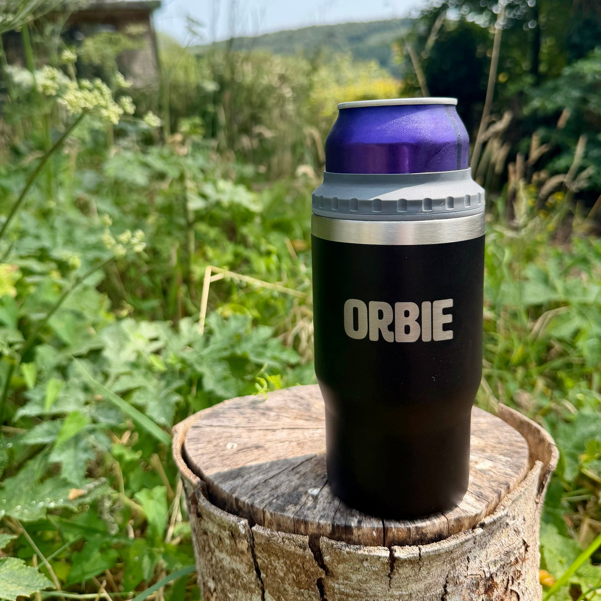 Black Orbie 12oz cup holding a 330ml can in the can cooler collar, standing on a tree stump outdoors
