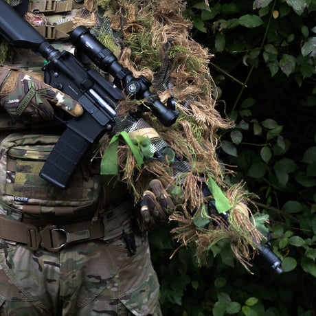 Zero Alpha Designs rifle ghillie ribbon wrapped around the handguard of a scoped rifle, hessian strands and green fabric leaf strips over a camouflage mesh strip