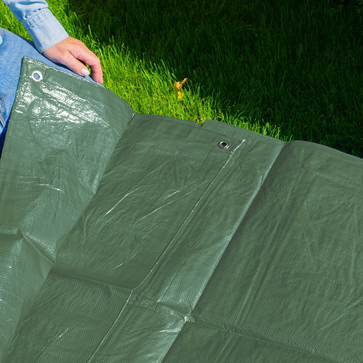Green M-Grade Tarpaulin 1m x 1m spread on a lawn with someone kneeling at the edge
