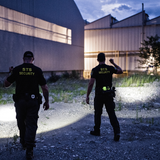 Two security officers walking an industrial site at dusk, lighting the ground with handheld torches
