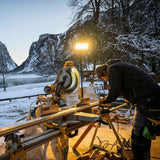 Joiner at a mitre saw on a snowy outdoor site at dusk, lit by a Ledlenser floodlight on a tall stand