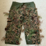 Zero Alpha Designs ghillie chaps laid flat, olive leg panels densely covered in hand-dyed hessian tufts and fabric leaves below the waistband