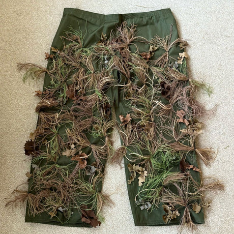 Zero Alpha Designs ghillie chaps laid flat, olive leg panels densely covered in hand-dyed hessian tufts and fabric leaves below the waistband