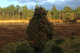 Rear view of the full cape ghillie suit dressed with bracken and conifer, standing in open heathland below a treeline