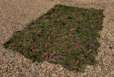 Zero Alpha Designs hide sheet laid flat on gravel at an angle, green tufts, tan strands and brown fabric leaves over a camouflage-printed base