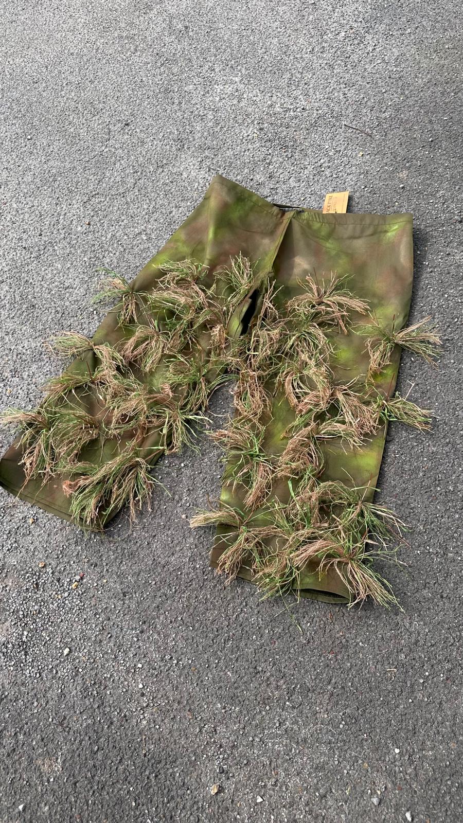 A lightly garnished pair of ghillie chaps, hessian tufts stitched across hand-sprayed green and brown leg panels with the Jack Pyke swing tag still attached