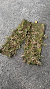 A lightly garnished pair of ghillie chaps, hessian tufts stitched across hand-sprayed green and brown leg panels with the Jack Pyke swing tag still attached