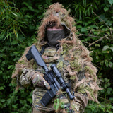 Head and shoulders ghillie suit worn front-on in hedgerow, tan and green hessian hanging clear of the rifle and chest rig