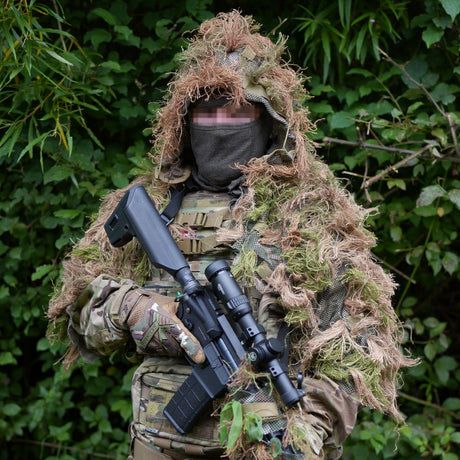 Head and shoulders ghillie suit worn front-on in hedgerow, tan and green hessian hanging clear of the rifle and chest rig