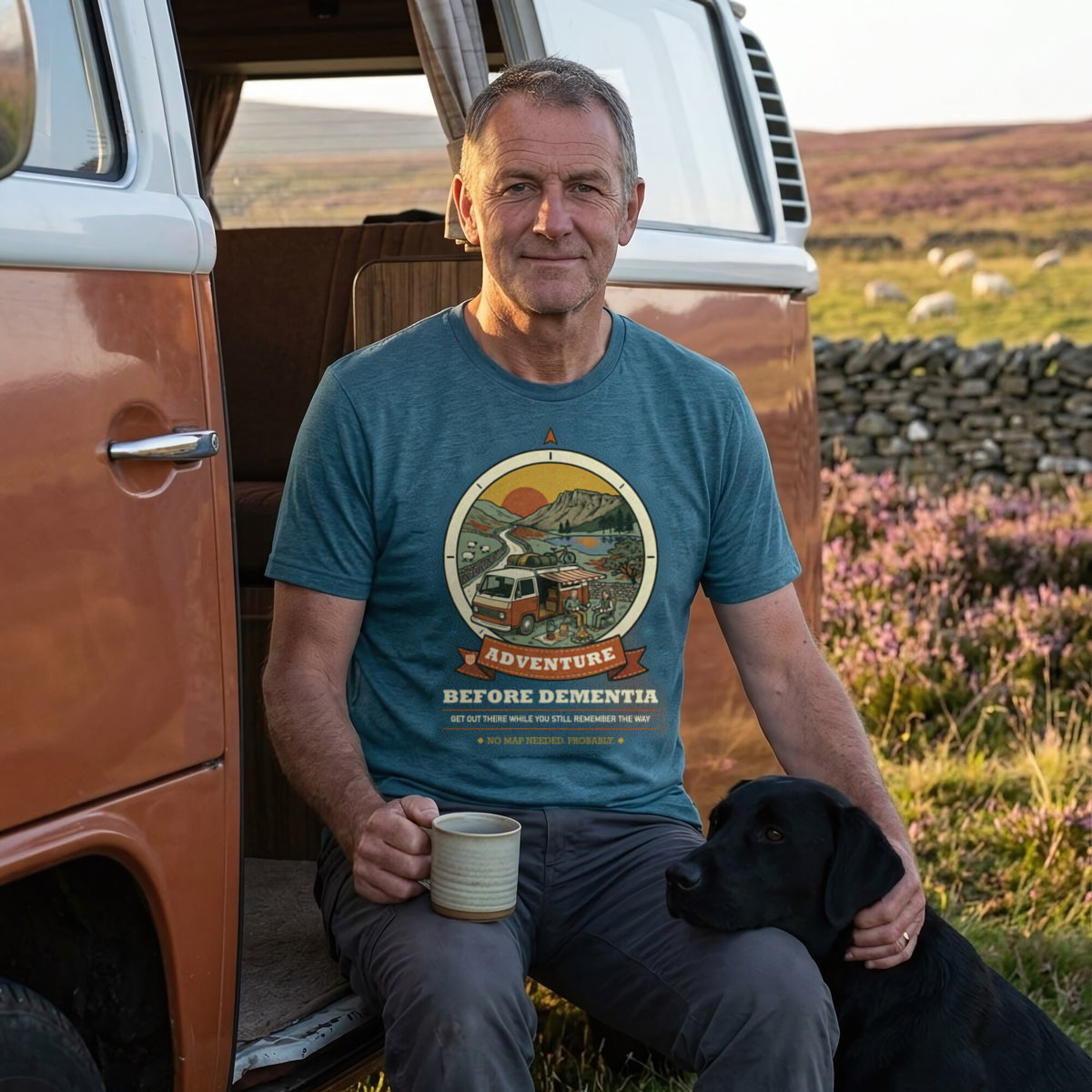Man in the Adventure Before Dementia Campervan T-Shirt in Heather Deep Teal sitting in a campervan doorway with a mug and his black Labrador on heather moorland
