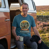 Man in the Adventure Before Dementia Campervan T-Shirt in Heather Deep Teal sitting in a campervan doorway with a mug and his black Labrador on heather moorland