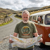 Bearded man in the Adventure Before Dementia Campervan T-Shirt in Military Green puzzling over a road map on a hill lane while his wife laughs and points from the campervan