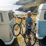 Smiling woman in the Adventure Before Dementia Campervan T-Shirt in Steel Blue lifting a bike off a campervan rack on a sunny clifftop
