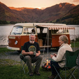 Man in the Adventure Before Dementia Campervan T-Shirt in Vintage Black laughing with his wife in camping chairs by a campfire, their campervan parked by a lake at dusk