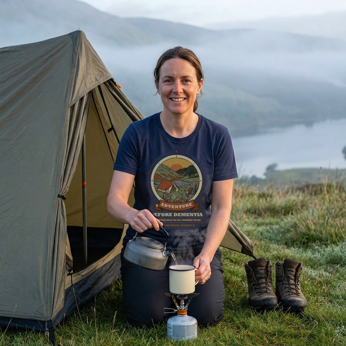 Woman in the Adventure Before Dementia Camping T-Shirt in Navy pouring a brew from a camping stove outside her tent at dawn